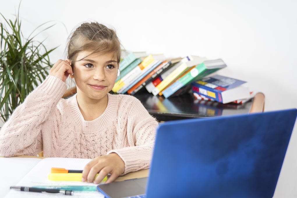 Ein Mädchen mit Kopfhörer sitzt vor einem Laptop, dahinter ein Stapel Schulbücher